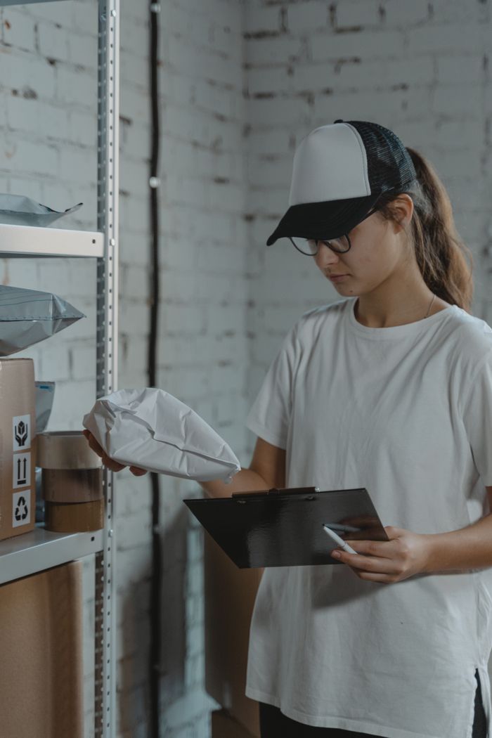 Young woman in white shirt and cap organizing packages in warehouse setting with clipboard.